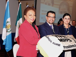 Three people standing together with a folded piece of cloth showing the MIRPS logo with "Presidencia" written on it - behind them are some of the flags of the MIRPS States