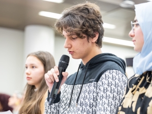 Three youths sitting in a row with other people listening behind them. The one in the middle is speaking into a microphone