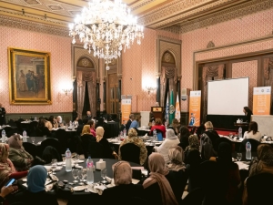 A meeting in what looks like an event room of a grand hotel - there are six round tables around which people are sitting, a stage with a Podium where someone is speaking, and a chandelier coming from the ceiling.