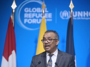 A man standing at a lectern speaking, there are flags behind him with the Global Refugee Forum logo on the wall behind