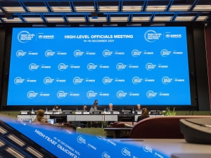 A view from the back of the room towards the stage, where there is a large screen with the High-Level Officials Meeting logo repeated across it. This, and the ceiling, are reflected in the foreground of the image, where a laptop or something is sitting