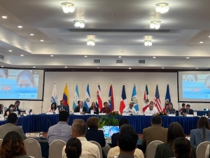 A meeting, with participants sitting behind tables laid out in a rectangle, with screens and flags behind the panel