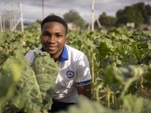 A young man crouching among green plants, smiling at the camera