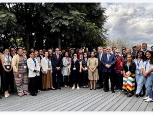A group photo of around 50 people standing on some wooden decking. There is a tree directly behind those to the left, and behind those on the right is a cloudy, but bright sky.