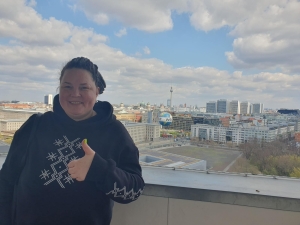 Anastasiia standing on the balcony of German Federal Ministry for Economic Cooperation and Development, behind her is the Berlin skyline, with the TV Tower (Fernsehturm) and Berlin Cathedral (Berliner Dom) in the distance and the Welt hot air balloon in the foreground. The sky is blue with white/light grey clouds.