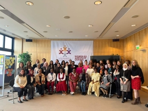A group photo of participants at the Action Network on Forced Displacement Annual Meeting - around 40 people, those at the front sitting on chairs with others standing behind them. Behind them on a wall is a large banner with the Action Network's logo and the words "Women as Agents of Change"