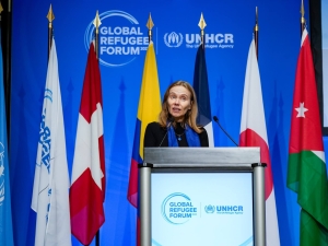 A woman standing at a podium with the Global Refugee Forum logo on it. Behind her are flags on poles, and behind those a blue backdrop with the Global Refugee Forum logo on it.