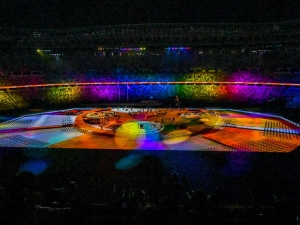 Inside the stadium, it is dark, except for the centre which is lit up with a lot of different coloured bright lights. In the middle, there are people on the stage, and the stands are filled with crowds