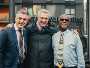 Three men standing together outside on what looks like a street in the centre of a town. The man in the middle has his arms around the shoulders of the other two. They are all smiling