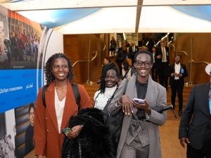 Four people walking through the entry tunnel - an indoor structure within the conference centre with images and the word "Welcome" in different languages on the wall and a tented roof. The four people are smiling at the camera. Behind them more people are descending a staircase coming towards the walk way