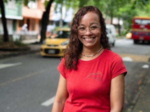 A woman standing in a tree-lined street, smiling at the camera. Behind her, there is a car driving one way and a bus driving in the other direction