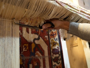 A rug being woven - it is hanging down, and a hand is holding scissors trimming the threads