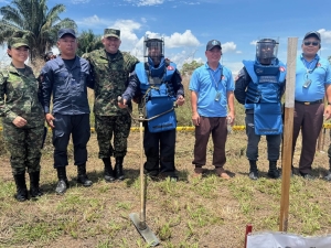 Eight people standing side by side in a field. They are dressed in a mixture of army fatigues and bomb-disposal type safety clothing, including helmets. They have tools, that look a bit like rakes, on the ground in front of them. The sky behind them is blue.