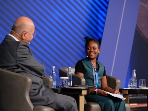The High Commissioner and Ms. Magesa sitting in chairs on stage. The photo is taken from the side, showing the side and back of the High Commissioner, and the face of Ms. Magesa. She is smiling.