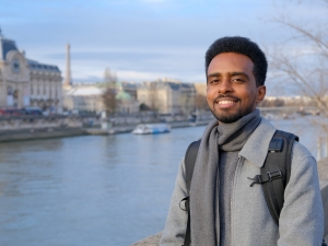 A man standing next to a river. He is smiling at the camera. On the other side of the river are some grand buildings, and in the distance is a boat.