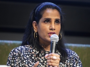 Ruven speaking into a microphone. A close up image only showing her upper body and torso, she is wearing a floral shirt, and holding out one hand to gesture while speaking.