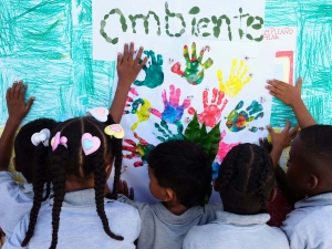 Children painting a picture with their hands. The photo is taken from behind the 5-6 children, and they have made handprints in different colours on a piece of paper, with the word 'ambiente' painted in what looks like fingerprints.