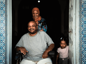 Family in a doorway, man is in a wheelchair with woman standing behind him. They are both laughing. Next to them, a little girl frowns at the camera