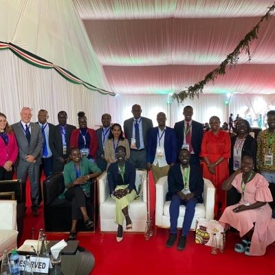 A group photo of refugee participants and UNHCR staff. Some are sitting in armchairs, others standing behind, all smiling and looking at the camera