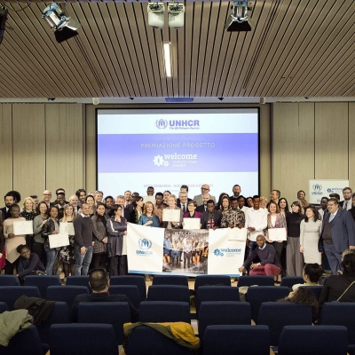 A large group photo from an event - they are standing on stage, holding a large Welcome banner with a screen behind them advertising the welcome project