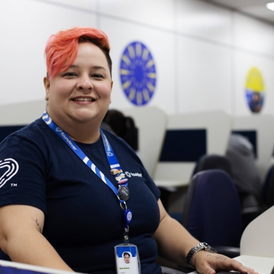 A woman sitting at a desk. She has a laptop in front of her, but is turning slightly sideways to look at the camera. She is smiling. Her hair is dyed bright orange and she is wearing a lanyard round her neck.