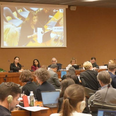 Photo from the event: a panel of five people sit facing the audience, while they listen to someone in the audience speaking. The person speaking is shown on a screen behind the panel.