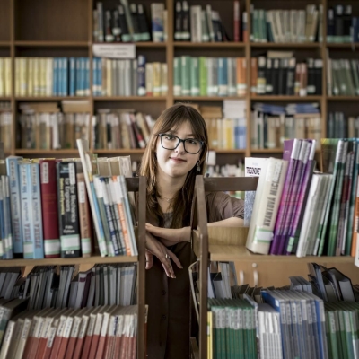 Olha standing in a library, leaning between two bookcases, looking at the camera