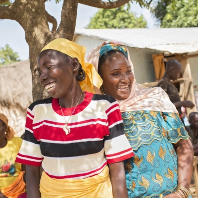Two women standing together under a tree laughing. There are other people and buildings in the background