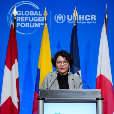 A woman speaking at a podium with eight different flags behind her and the Global Refugee Forum logo on the wall behind the flags