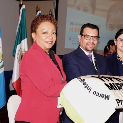 Three people standing together with a folded piece of cloth showing the MIRPS logo with "Presidencia" written on it - behind them are some of the flags of the MIRPS States