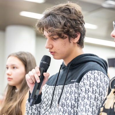 Three youths sitting in a row with other people listening behind them. The one in the middle is speaking into a microphone