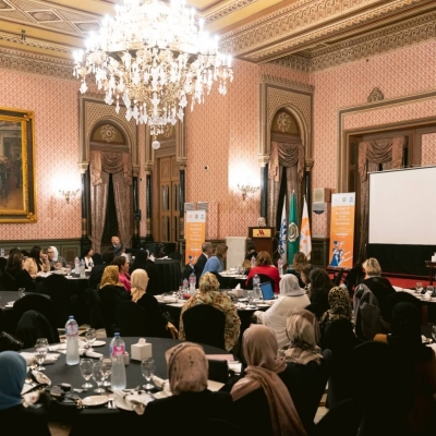 A meeting in what looks like an event room of a grand hotel - there are six round tables around which people are sitting, a stage with a Podium where someone is speaking, and a chandelier coming from the ceiling.