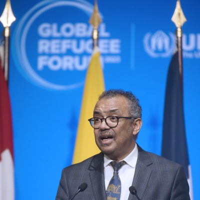 A man standing at a lectern speaking, there are flags behind him with the Global Refugee Forum logo on the wall behind