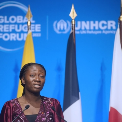 A woman standing at a podium speaking into microphones. Behind her are five flags, and behind the flags a blue background with the Global Refugee Forum logo on it