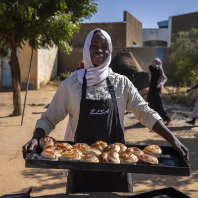 A woman standing holding a tray of baked swirls (cinnamon or raisin type of pastry) - she is wearing an apron, smiling at the camera, standing outside with a tree and some buildings in the background as two people walk behind her