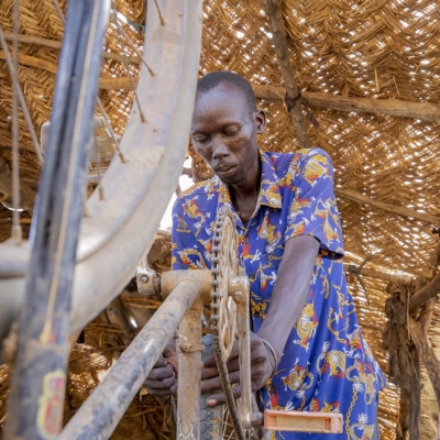 A man is repairing a bike, which is turned upside down. In the foreground is one of the bikes wheels, with the pedals and cogs visible behind, further back, the man is leaning over the back wheel of the bike, holding it with one hand and what looks to be a screwdriver in the other. Overhead there is a thatched roof held up with large logs