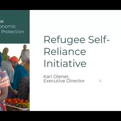A screenshot from the webinar showing a slide from the presentation, with the name of the multi-stakeholder pledge, the words "Refugee Self-Reliance Initiative- Kari Diener, Executive Director", a photo of a woman smiling in a market, three of the videos of participants in the call and a counter showing another 82 other people are in it too.
