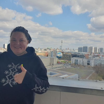 Anastasiia standing on the balcony of German Federal Ministry for Economic Cooperation and Development, behind her is the Berlin skyline, with the TV Tower (Fernsehturm) and Berlin Cathedral (Berliner Dom) in the distance and the Welt hot air balloon in the foreground. The sky is blue with white/light grey clouds.