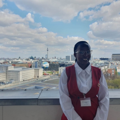 Rebecca standing on the balcony of German Federal Ministry for Economic Cooperation and Development, behind her is the Berlin skyline, with the TV Tower (Fernsehturm) and Berlin Cathedral (Berliner Dom) in the distance and the Welt hot air balloon in the foreground. The sky is blue with white/light grey clouds.