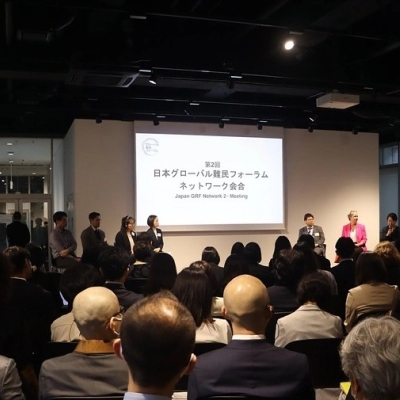A photo from the meeting, taken at the back of the room, showing participants sitting down, with eight people standing at the front facing them, the lights shining on them, and a screen behind them saying with some writing in Japanese, and in English the words "Japan GRF Network 2nd Meeting"