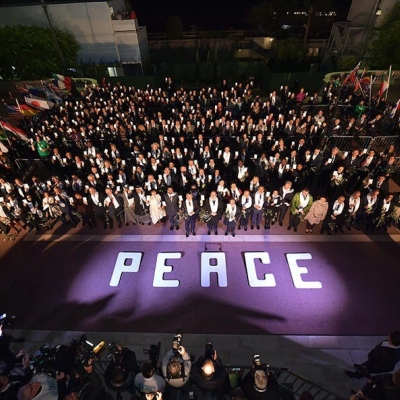 An aerial view of a large group of people standing outside, with a purple carpet and word peace written on the ground in front of them. There are flags near the people, and on the other side of the carpet are photographers and videographers