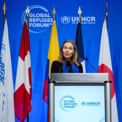 A woman standing at a podium with the Global Refugee Forum logo on it. Behind her are flags on poles, and behind those a blue backdrop with the Global Refugee Forum logo on it.