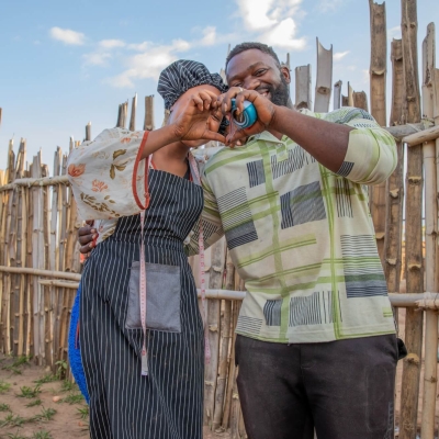 A man and a woman standing together outside, with a wooden fence behind them. They have their arms around one another, and are making a heart shape with his left hand and her right.