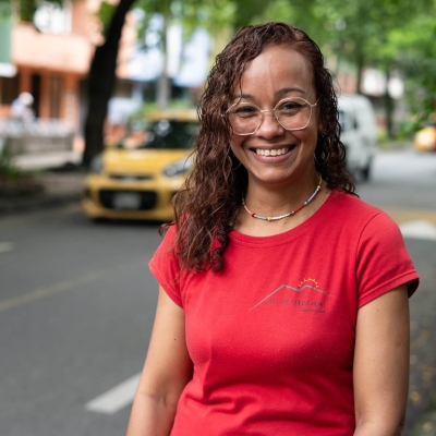 A woman standing in a tree-lined street, smiling at the camera. Behind her, there is a car driving one way and a bus driving in the other direction