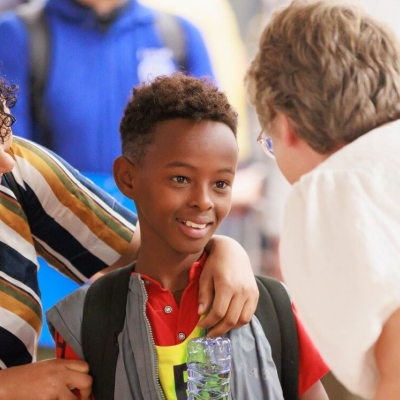 A boy being greeted by a woman, who is bending down to speak to him. He is holding a bottle of water upside down, smiling, while his mother has her arm around his shoulders and is looking at him
