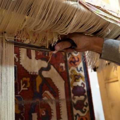 A rug being woven - it is hanging down, and a hand is holding scissors trimming the threads