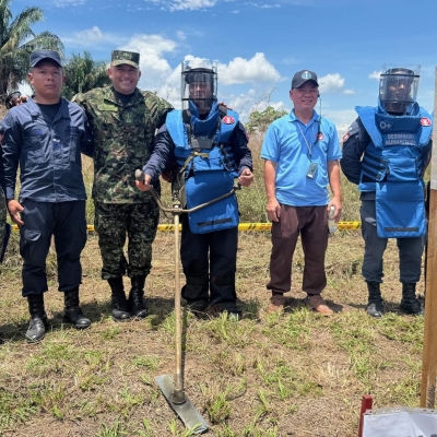 Eight people standing side by side in a field. They are dressed in a mixture of army fatigues and bomb-disposal type safety clothing, including helmets. They have tools, that look a bit like rakes, on the ground in front of them. The sky behind them is blue.