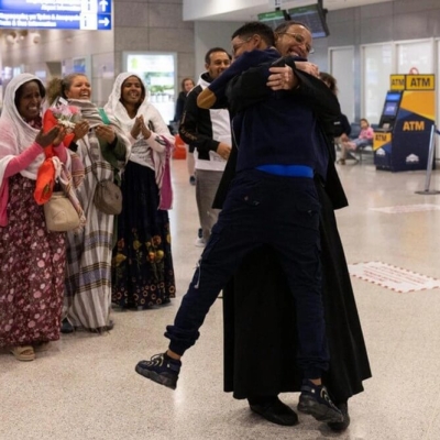 Father Solomon embraces tightly his youngest son, Yoftahie, who has just landed to Athens with his mother and siblings under the gaze of female members of the Eritrean Orthodox congregation in Greece, who joined Father Solomon welcoming his family at the International Airport of Athens