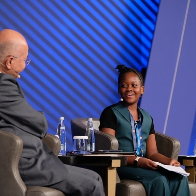 The High Commissioner and Ms. Magesa sitting in chairs on stage. The photo is taken from the side, showing the side and back of the High Commissioner, and the face of Ms. Magesa. She is smiling.