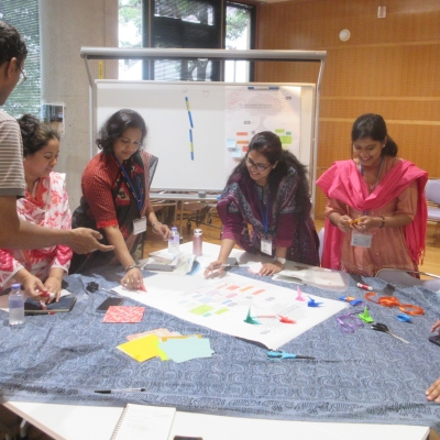 A group of six people standing round a table putting colourful pieces of paper (perhaps some origami?) on a large sheet of paper